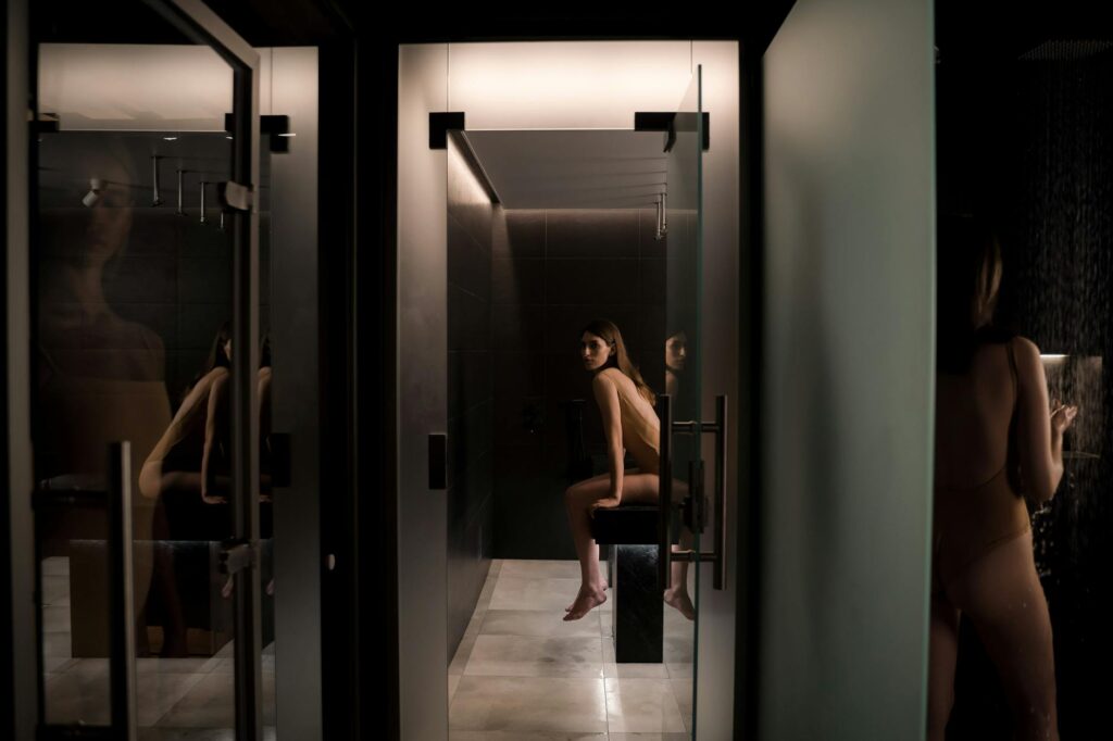 young woman taking a shower by the doorway of spa sauna where her friend is waiting on the massage table